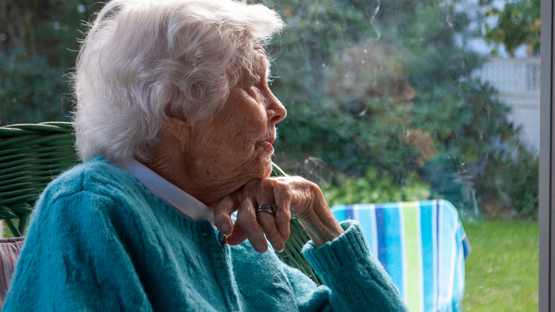 Elderly woman in a teal sweater gazing thoughtfully out a window.