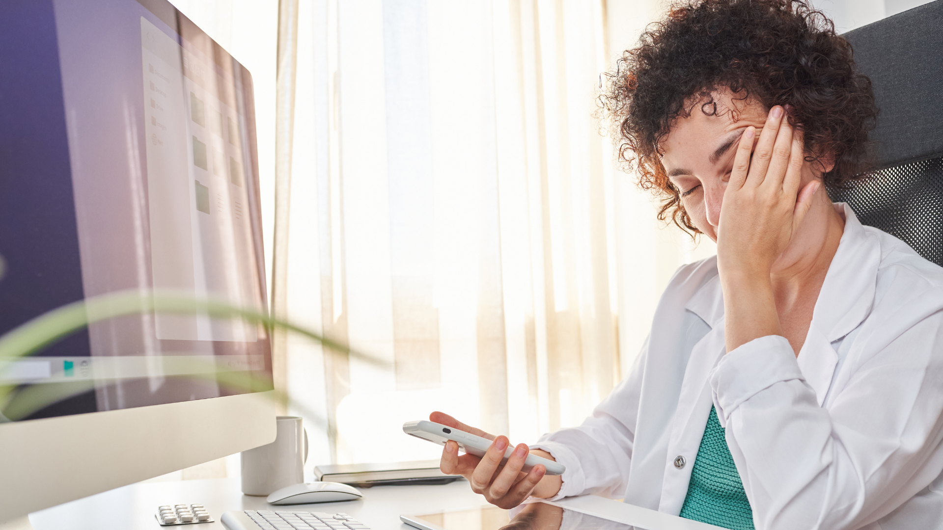 Exhausted doctor holding a phone, sitting at her desk with eyes closed in frustration.